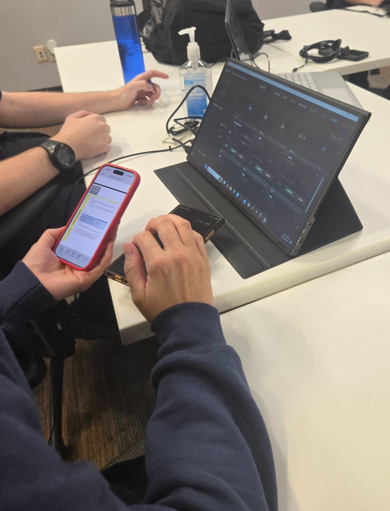 Two medics at a table. One holds a phone open to a Levophed reference while a portable monitor runs the simulator.
